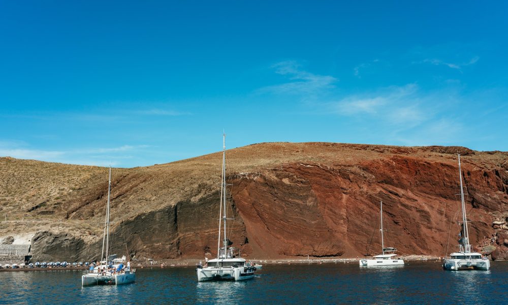Catamarans anchored at the Red Beach, one of the popular stops on our Joy Daytime Santorini Half Day Cruise by Spiridakos Sailing Cruises.