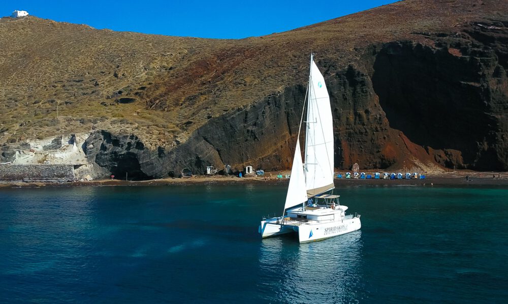 Spiridakos Sailing Cruises catamaran pictured against the Red Rock beach, one of the landmark stops during a Santorini caldera cruise.