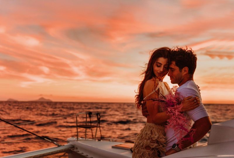 Couple embrace on a vessel at sea with the sunset sky background, enjoying a romantic dinner in Santorini. 