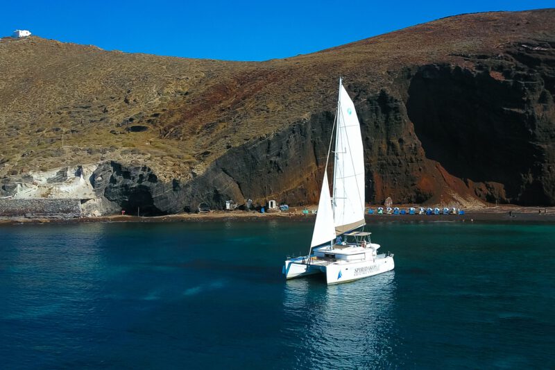 Spiridakos Sailing Cruises catamaran pictured against the Red Rock beach, one of the landmark stops during a Santorini caldera cruise.